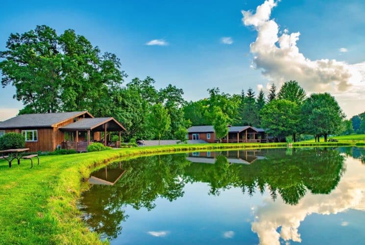 Exterior view of a property with brown wooden buildings nestled next to a small pond surrounded by large green trees