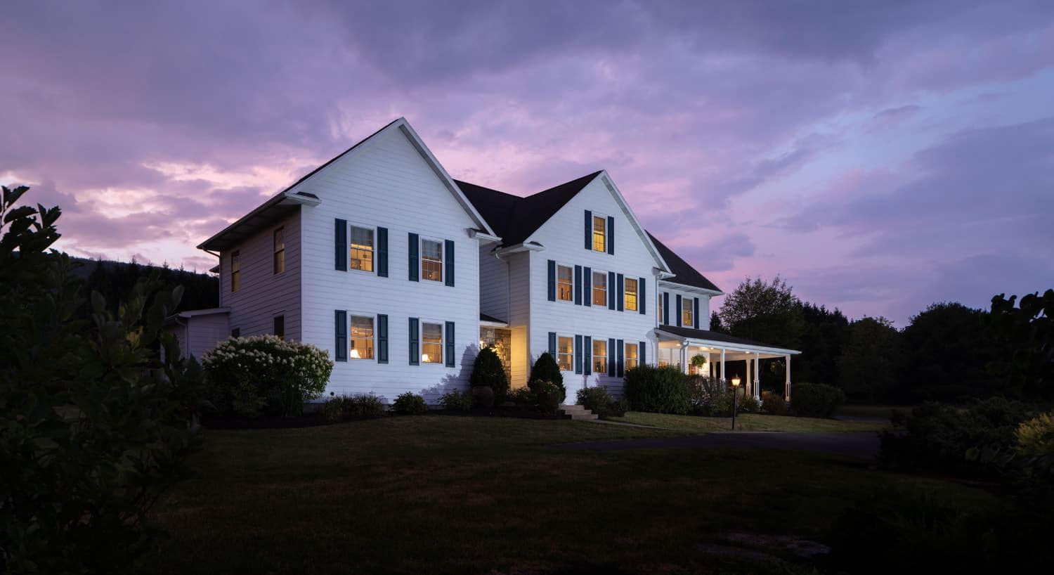 Exterior view of the property painted white with navy shutters and surrounded by green grass, bushes, and trees at dusk