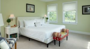 Bedroom with light green walls, carpeting, white upholstered bed, white bedding, white nightstands with lamps, and colorful footstool