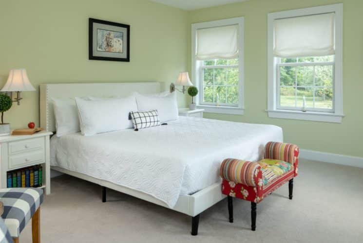 Bedroom with light green walls, carpeting, white upholstered bed, white bedding, white nightstands with lamps, and colorful footstool