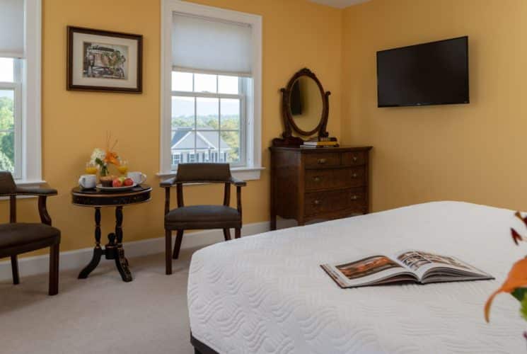 Bedroom with light orange walls, carpeting, white bedding, sitting area, wooden dresser, and wall-mounted TV