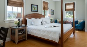 Bedroom with light colored walls, hardwood flooring, wooden four-poster bed, white bedding, wooden nightstands with lamps, and sitting area