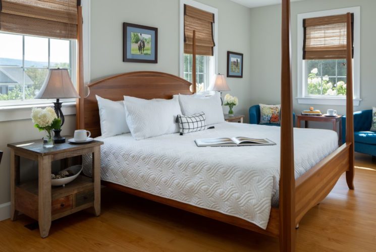 Bedroom with light colored walls, hardwood flooring, wooden four-poster bed, white bedding, wooden nightstands with lamps, and sitting area