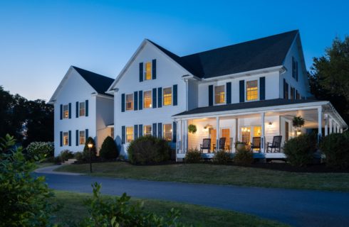 Exterior view of the property painted white with navy shutters and surrounded by green grass, bushes, and trees at dusk