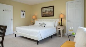 Bedroom with light yellow walls, carpeting, white upholstered bed, white bedding, and wooden nightstands with lamps