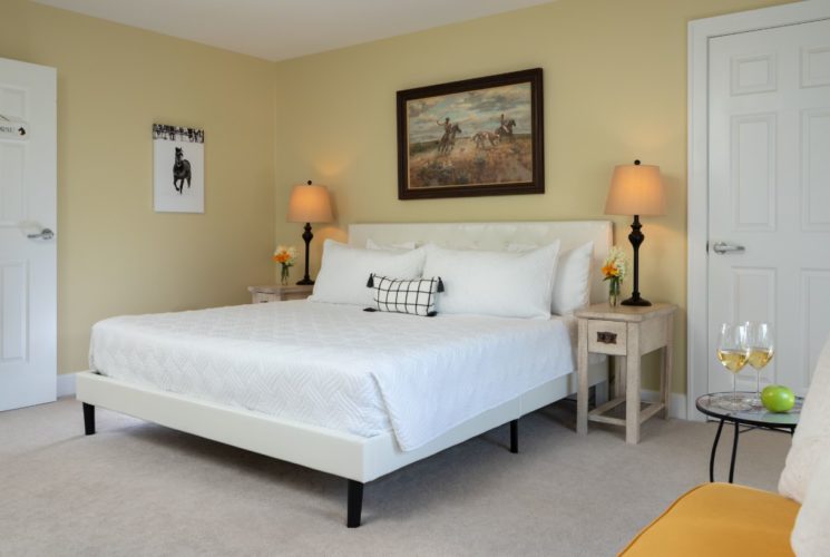 Bedroom with light yellow walls, carpeting, white upholstered bed, white bedding, and wooden nightstands with lamps