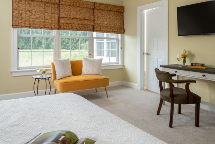 Bedroom with light yellow walls, white trim, white bedding, desk, wooden chair, orange love seat, and wall-mounted TV