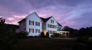 Exterior view of the property painted white with navy shutters and surrounded by green grass, bushes, and trees at dusk