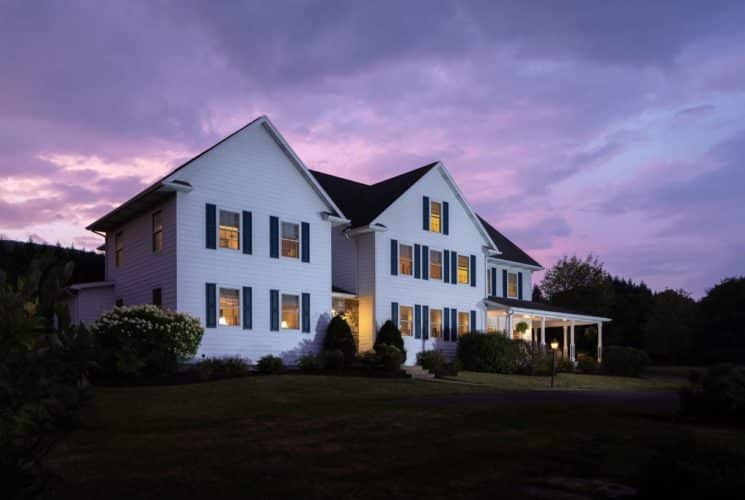 Exterior view of the property painted white with navy shutters and surrounded by green grass, bushes, and trees at dusk