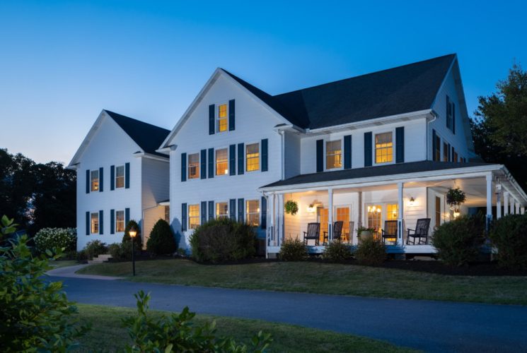 Exterior view of the property painted white with navy shutters and surrounded by green grass, bushes, and trees at dusk