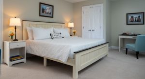 Bedroom with light colored walls, carpeting, light wooden bed, white bedding, white nightstands with lamps, and desk with chair