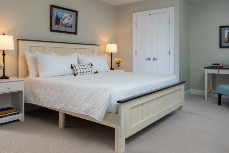 Bedroom with light colored walls, carpeting, light wooden bed, white bedding, white nightstands with lamps, and desk with chair