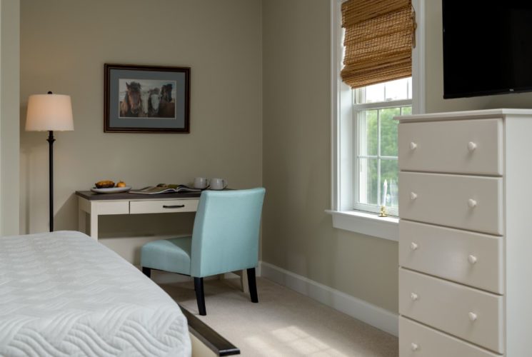 Bedroom with light colored walls, carpeting, white bedding, desk, light blue upholstered chair, and white dresser