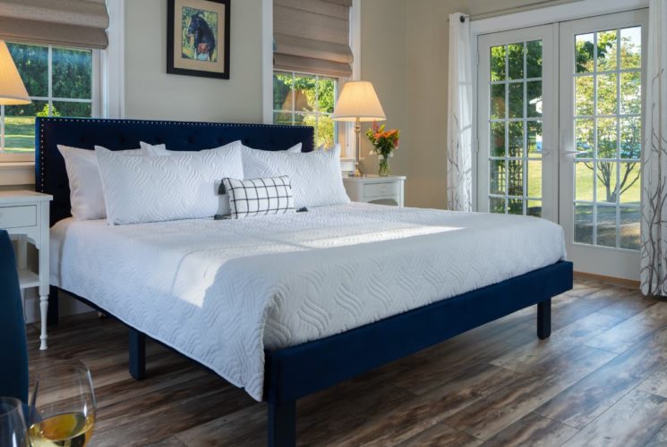 Bedroom with light colored walls, hardwood flooring, navy upholstered bed, white bedding, white nightstands with lamps, and views to the outside