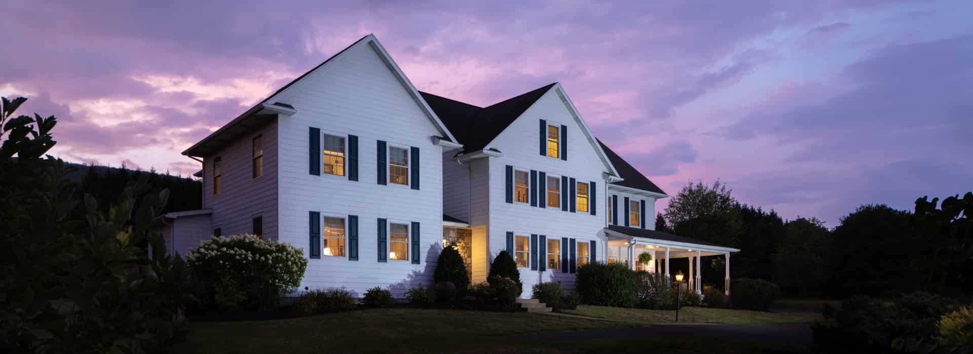 Exterior view of the property painted white with navy shutters and surrounded by green grass, bushes, and trees at dusk