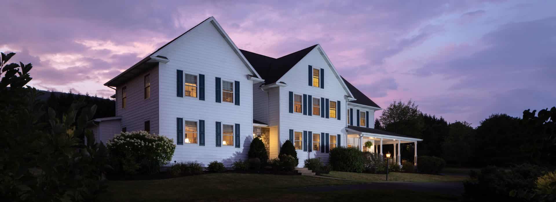 Exterior view of the property painted white with navy shutters and surrounded by green grass, bushes, and trees at dusk