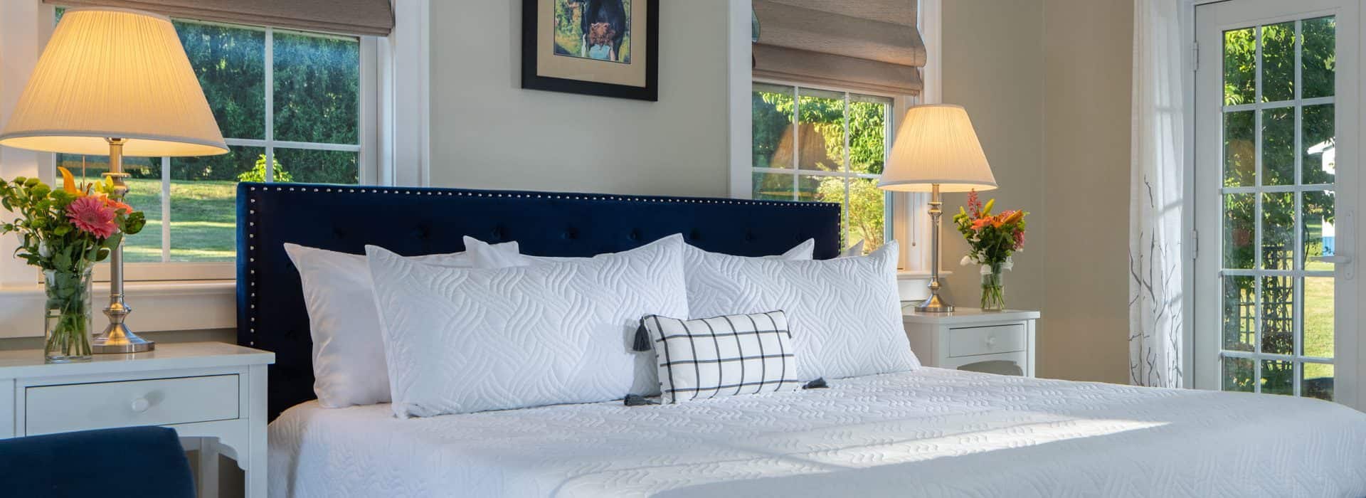 Close up view of navy upholstered bed, white bedding, white nightstands with lamps, and views to the outside