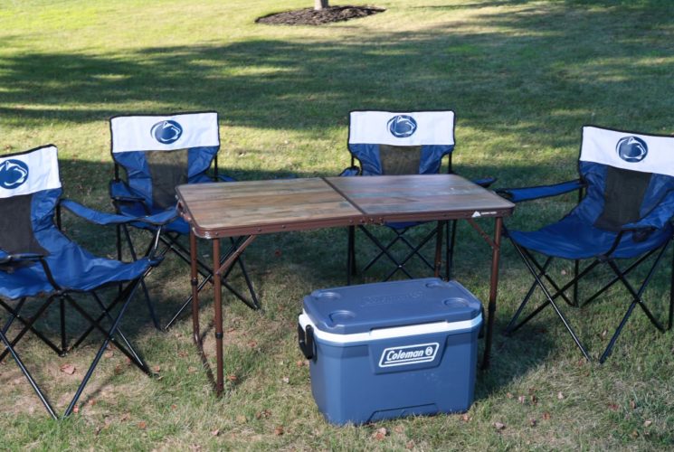 Four blue and black foldout chairs, brown folding table, and blue cooler on green grass