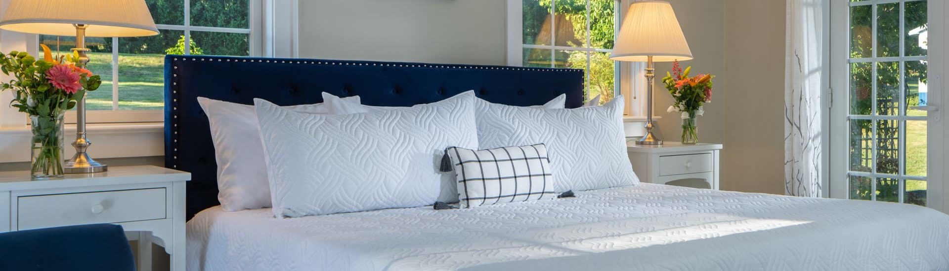 Close up view of navy upholstered bed, white bedding, wooden nightstands with lamps, and windows