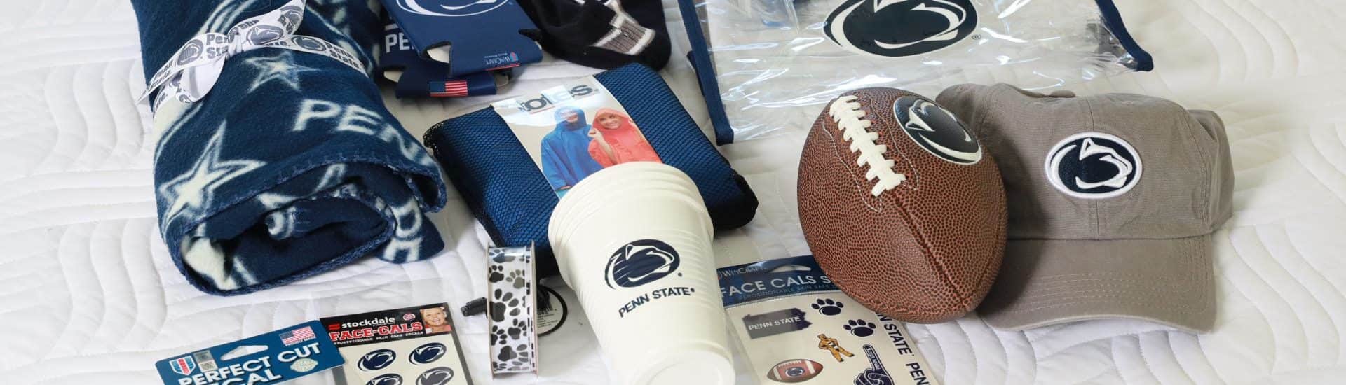 Penn State branded hat, football, cup, stickers, blanket, clear bag, and cozies on a white blanket