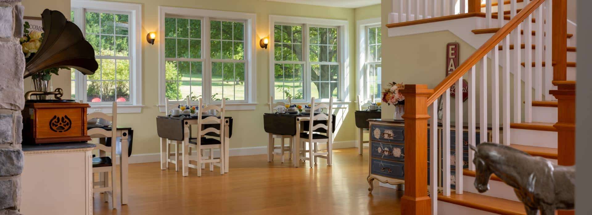 Dining room with light yellow walls, hardwood flooring, wooden tables and chairs, wooden staircase, and multiple windows with views to the outside