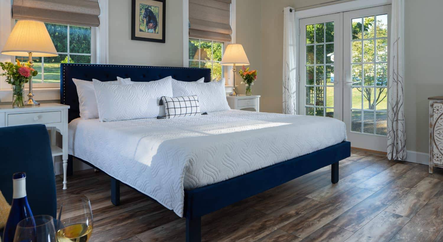 Bedroom with light colored walls, hardwood flooring, navy upholstered bed, white bedding, white nightstands with lamps, and views to the outside